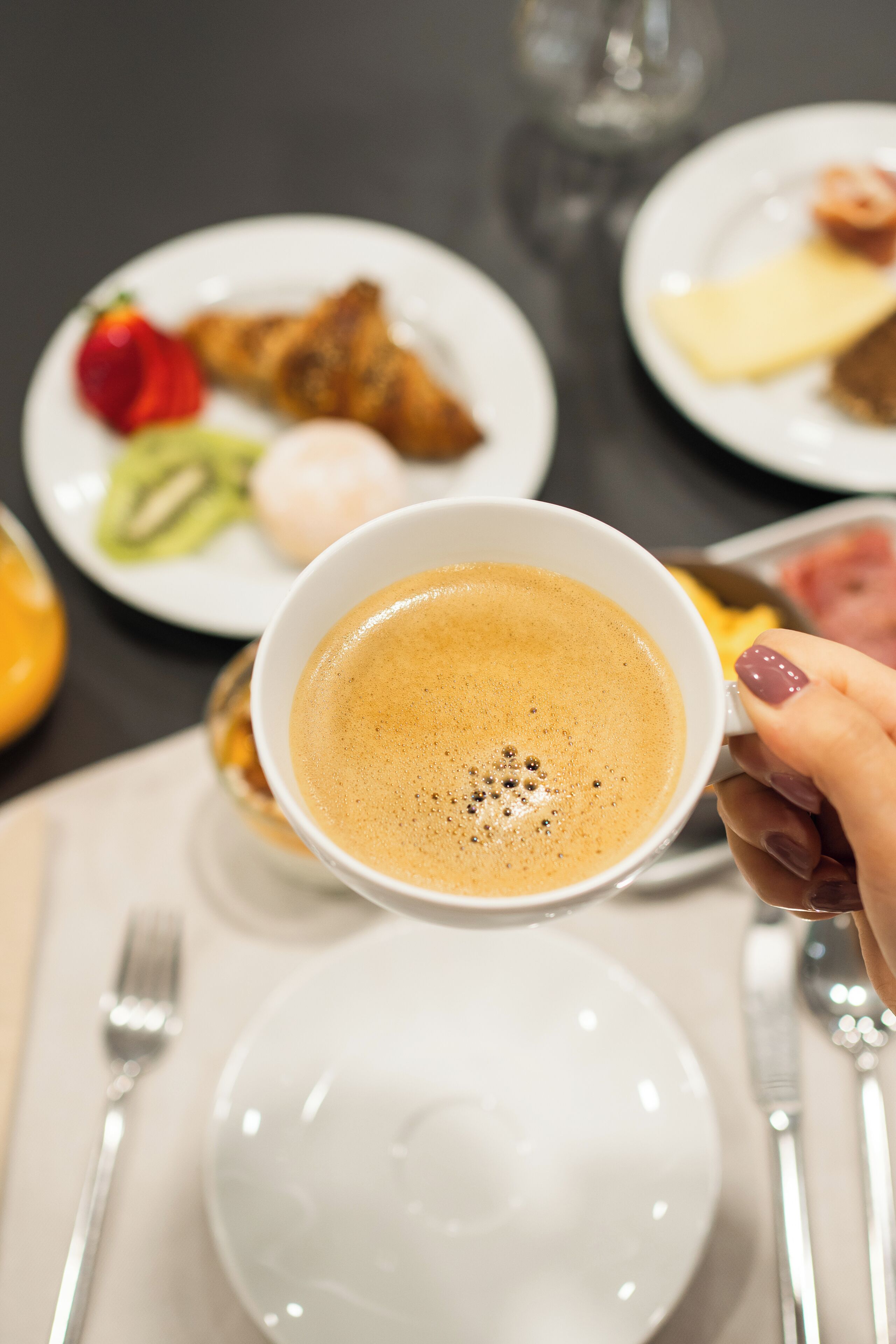 Una mano sosteniendo una taza de café, con un fondo borroso de una mesa de comedor con platos de comida.