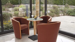 A modern outdoor seating area with two brown chairs and a glass table, surrounded by a stone wall and greenery. - Hotel Germania (Reutlingen)