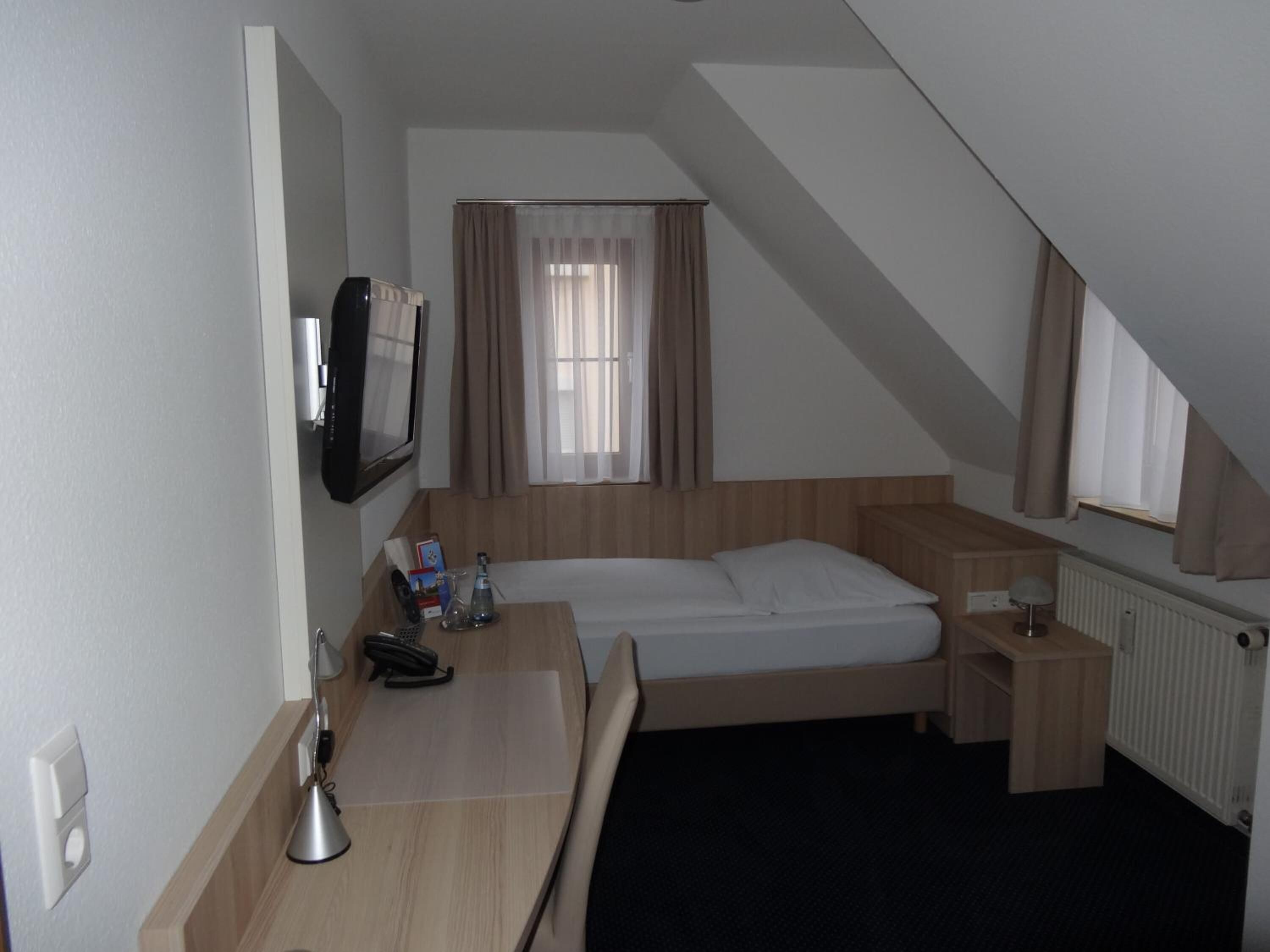 Een kleine, goed verlichte kamer met een bed, bureau, stoel, televisie en raam met gordijnen.