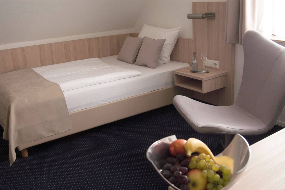 A hotel room with a single bed, a bedside table with a glass of water, a chair, and a bowl of fruit.
