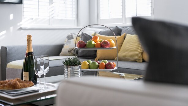 A dining area with a bottle of champagne, two glasses, a tray with croissants, and a fruit stand with apples and oranges. - Drayton (London)