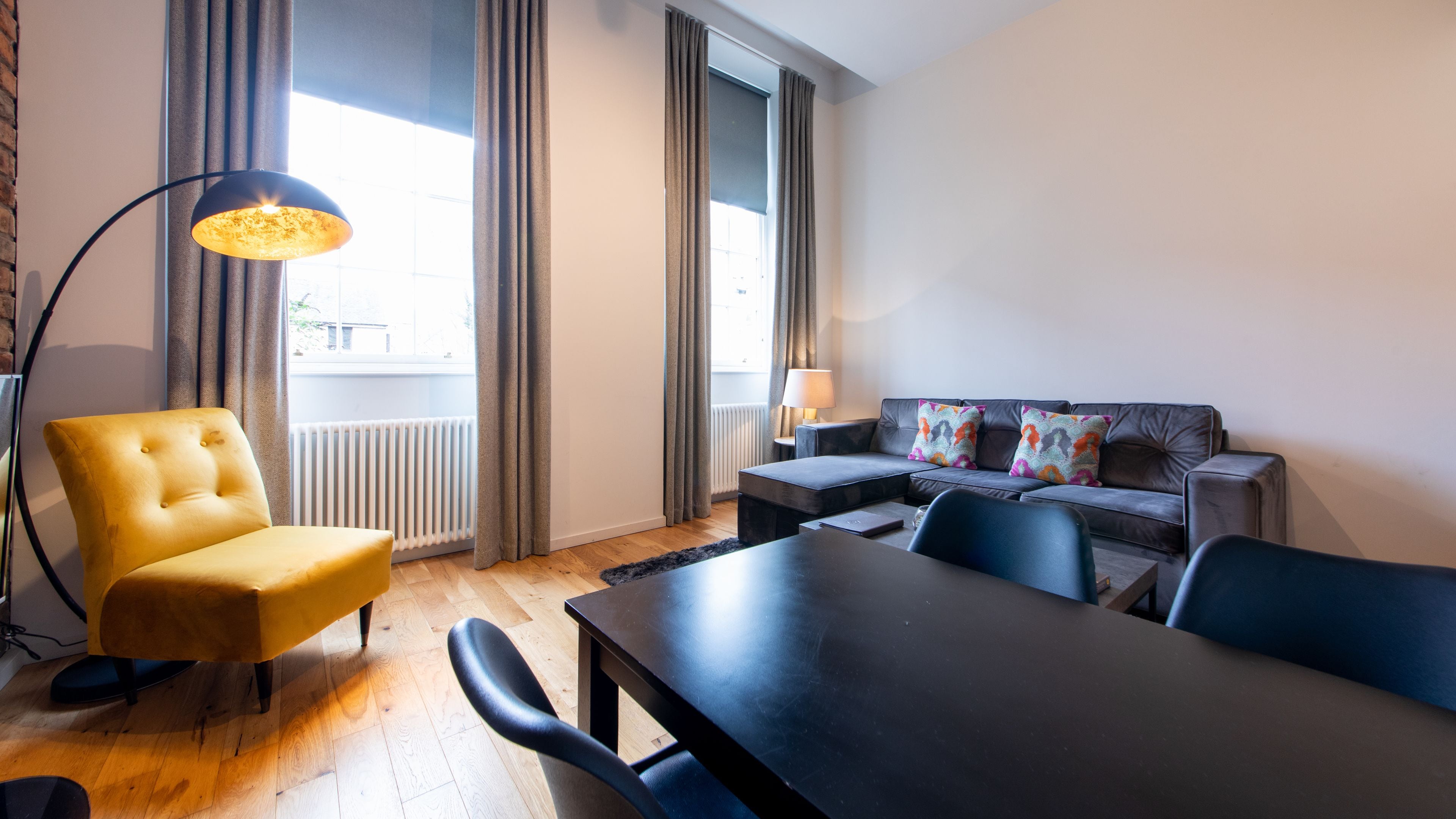 A modern living room with a yellow armchair, a black sofa with colorful cushions, a dining table, and a floor lamp.