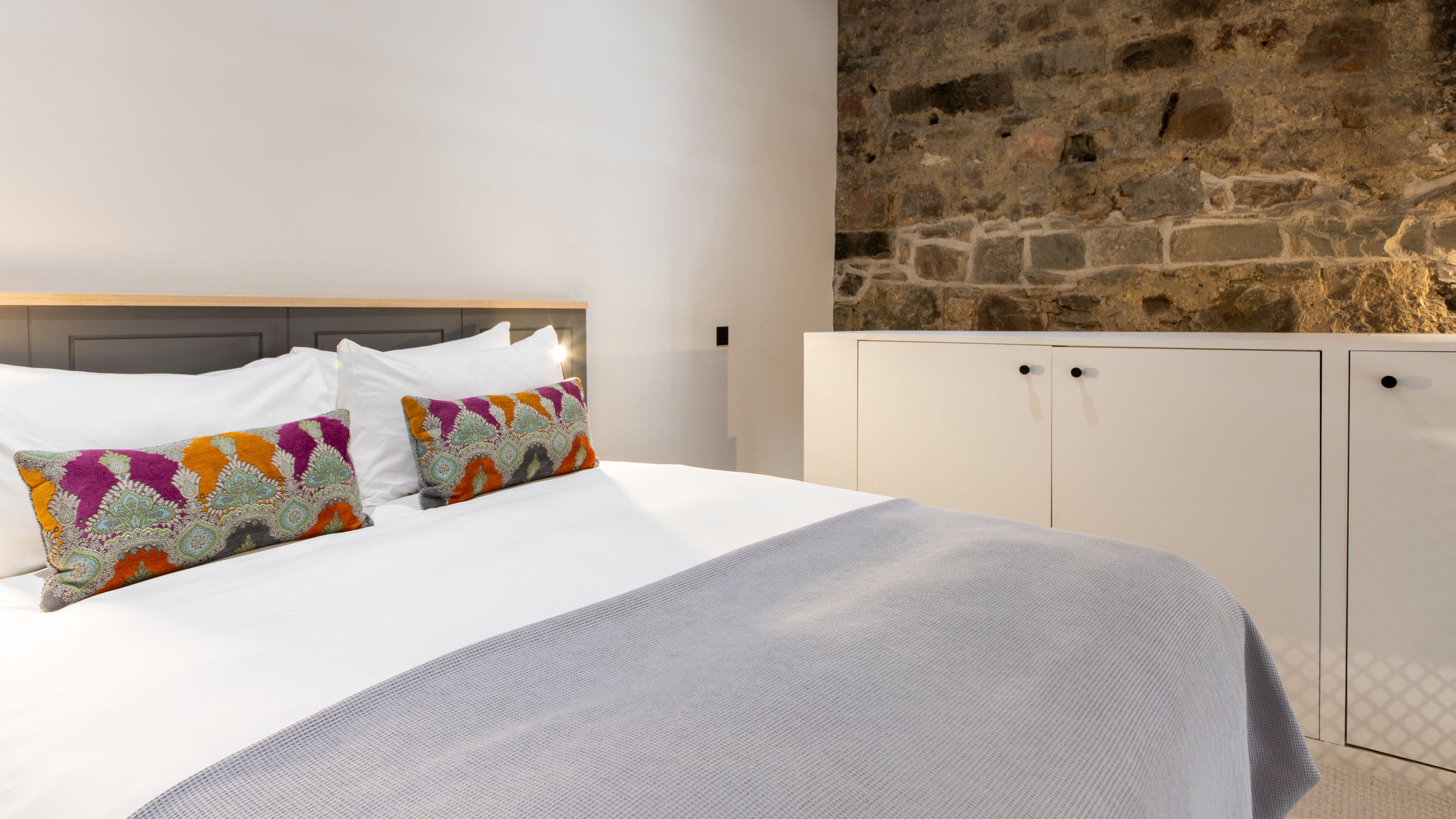 A bed with white linens and colorful patterned pillows, a stone wall, and white cabinets with black handles.