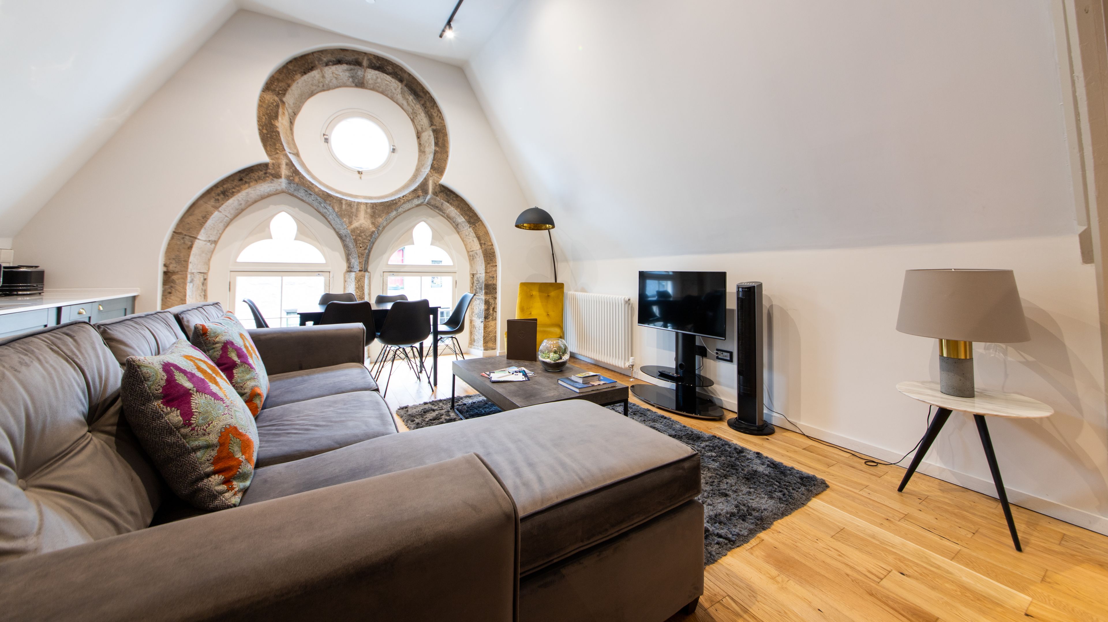 A modern living room with a grey sofa, a coffee table, and a TV. There is a dining area with chairs and a table in the background.