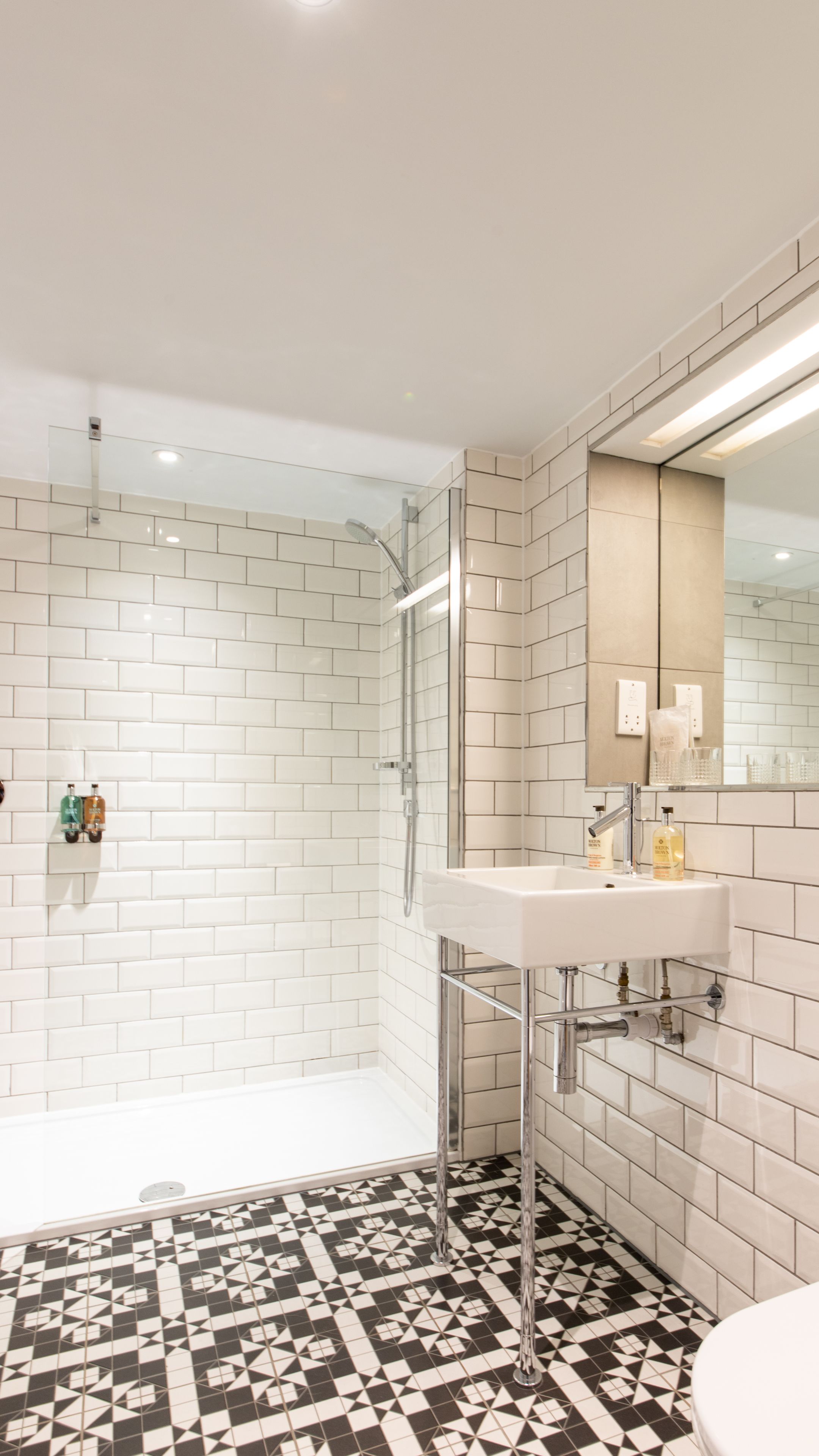 Une salle de bain moderne avec des murs recouverts de carreaux blancs, un sol à motifs noir et blanc, une douche en verre et un lavabo blanc avec un miroir au-dessus.