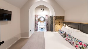 A bedroom with a large bed, a TV mounted on the wall, and an arched doorway leading to a bathroom. - Linton Collection - Blackfriars Lofts (Edinburgh)