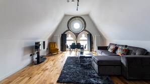 A modern loft with a high ceiling, wooden flooring, a grey sofa, a yellow armchair, and a dining area with a table and chairs. - Linton Collection - Blackfriars Lofts (Edinburgh)