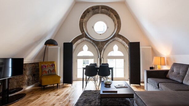 A modern loft with a high ceiling, exposed stone arches, a dining area with a table and chairs, a sofa, a TV, and a fireplace. - Linton Collection - Blackfriars Lofts (Edinburgh)