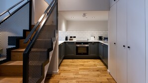 A modern kitchen with wooden flooring, a staircase with a metal railing, and white cabinets. - Linton Collection - Blackfriars Lofts (Edinburgh)
