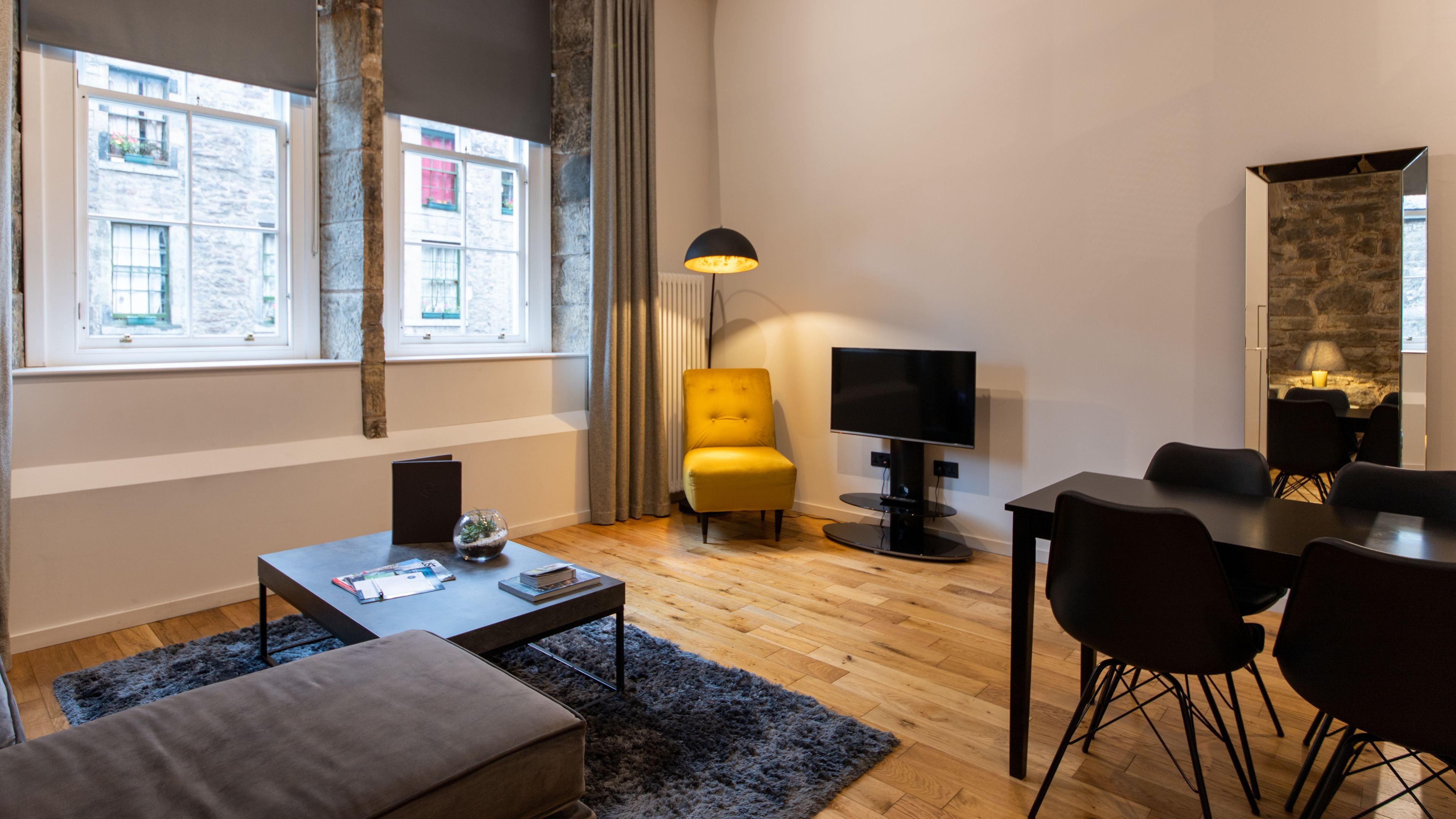 Una sala de estar moderna con un sofá, una mesa de centro y un sillón amarillo. Hay un televisor de pantalla plana montado en la pared, una mesa de comedor con sillas y una lámpara. Amplias ventanas ofrecen luz natural y vistas al exterior.