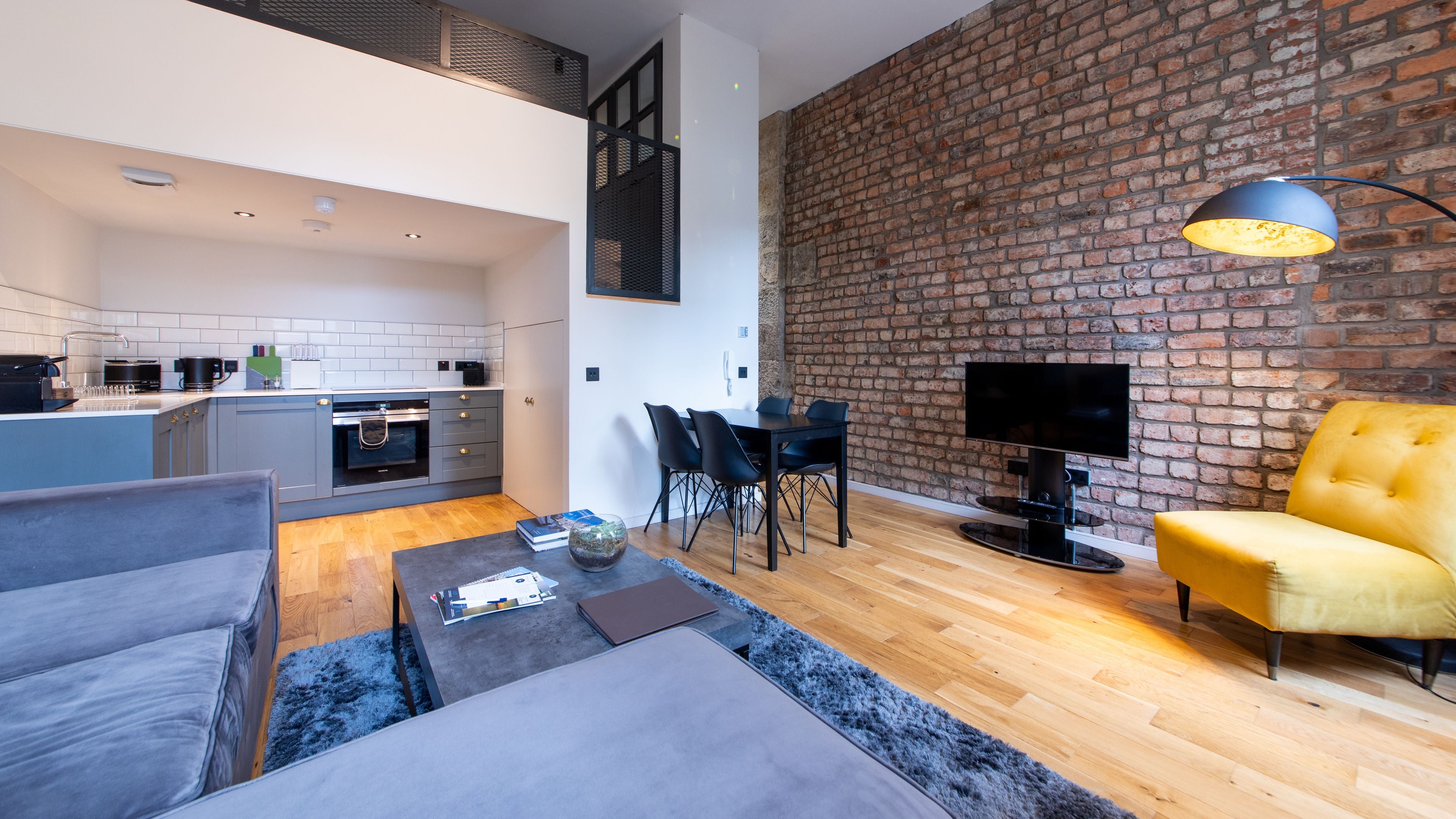 A modern living room with a sofa, coffee table, and a yellow armchair. A dining area with a table and chairs is adjacent to a kitchen with a brick wall. A flat-screen TV is mounted on the brick wall.