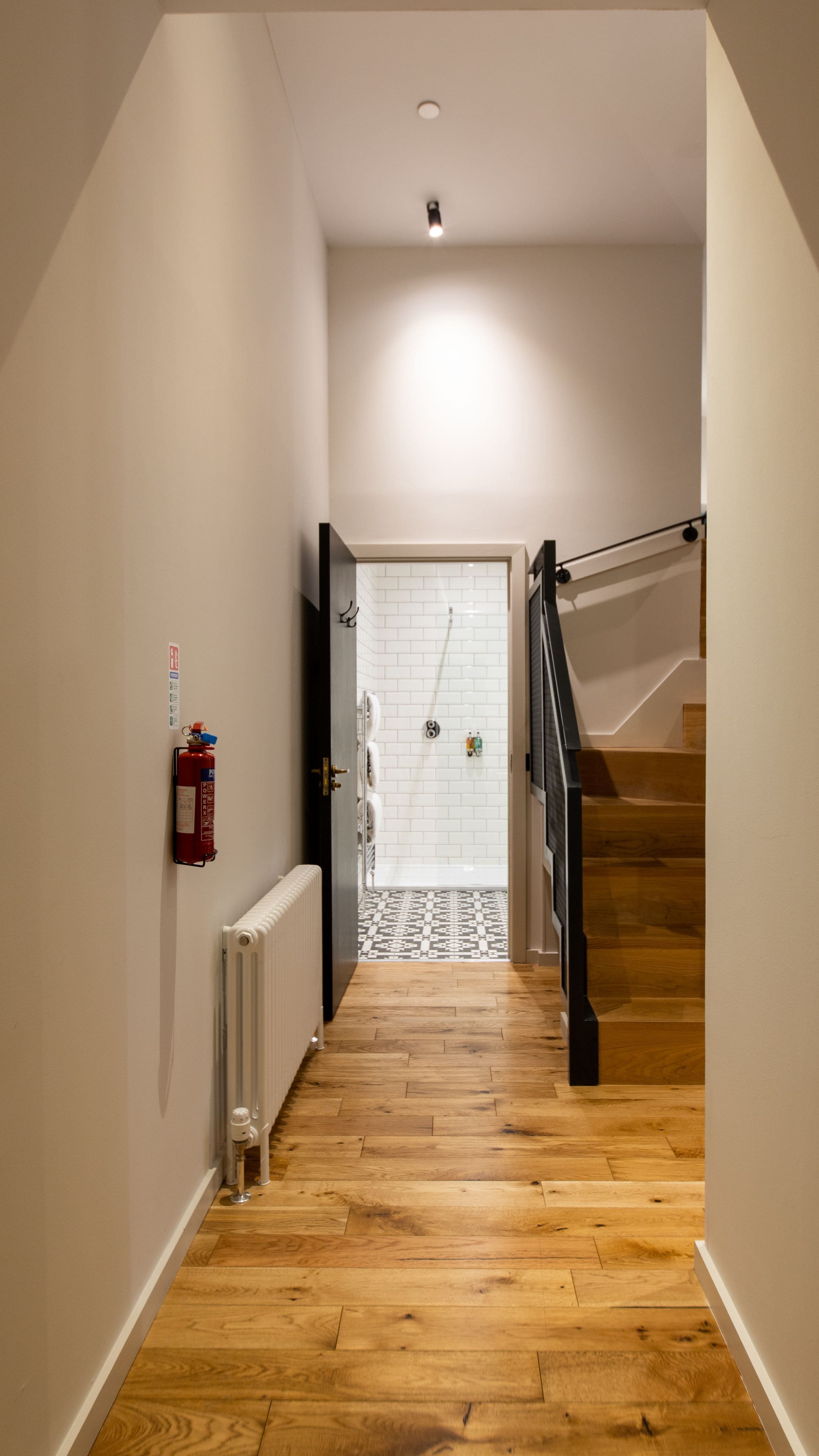A narrow hallway with wooden flooring, a white radiator, a fire extinguisher, and a tiled shower area at the end.
