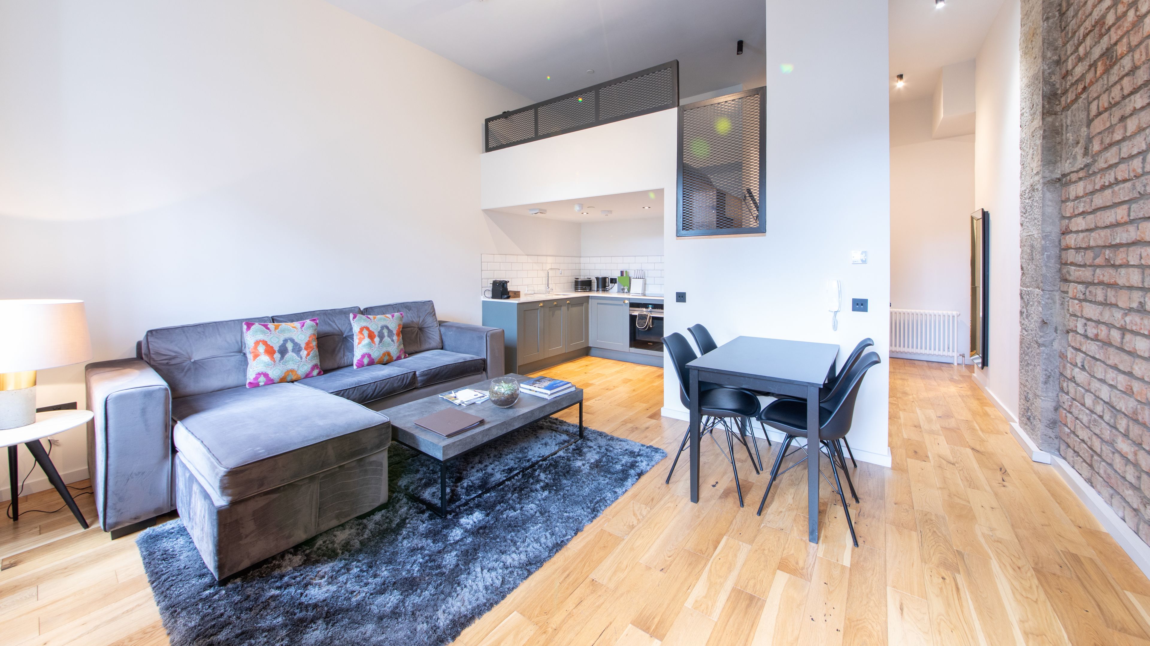Una sala moderna con un sofá gris, una mesa de centro y un comedor con mesa y sillas. Se ve una cocina al fondo.