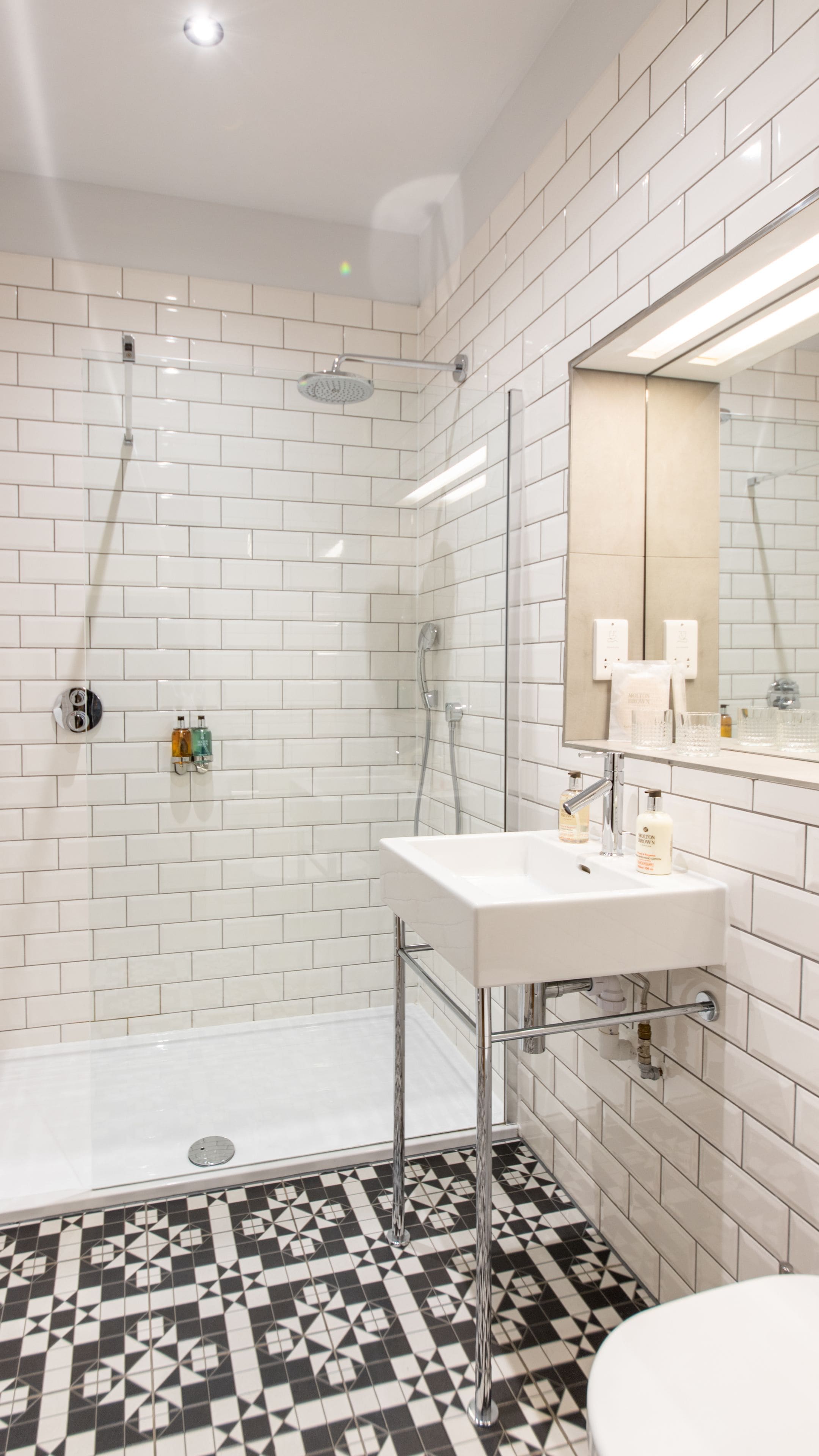 A modern bathroom with white subway tiles, a shower with a glass door, a white sink with chrome fixtures, and a patterned floor.
