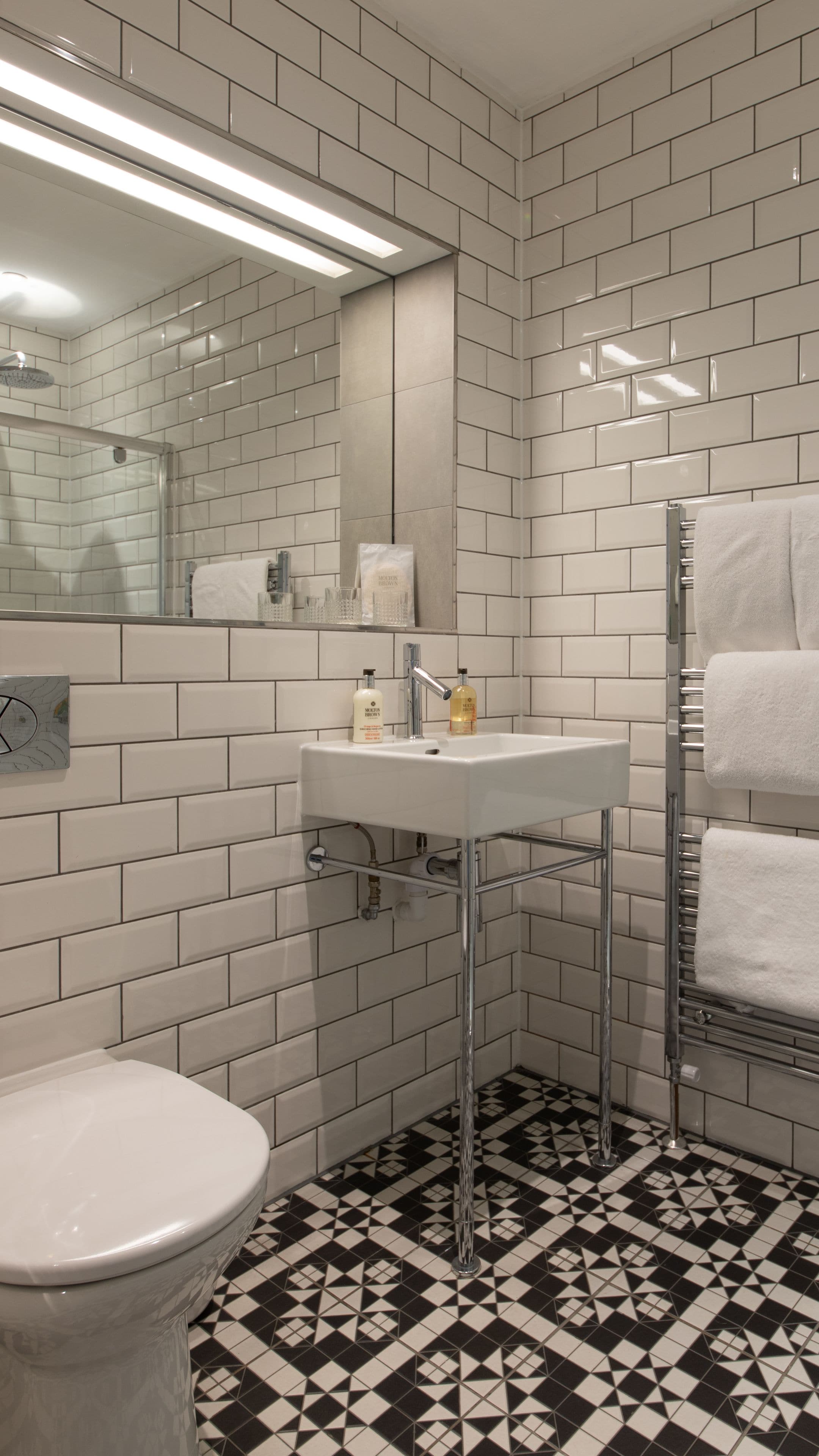 Une salle de bain avec des carreaux de céramique blancs rectangulaires, un lavabo blanc, un miroir, un porte-serviettes et des toilettes.
