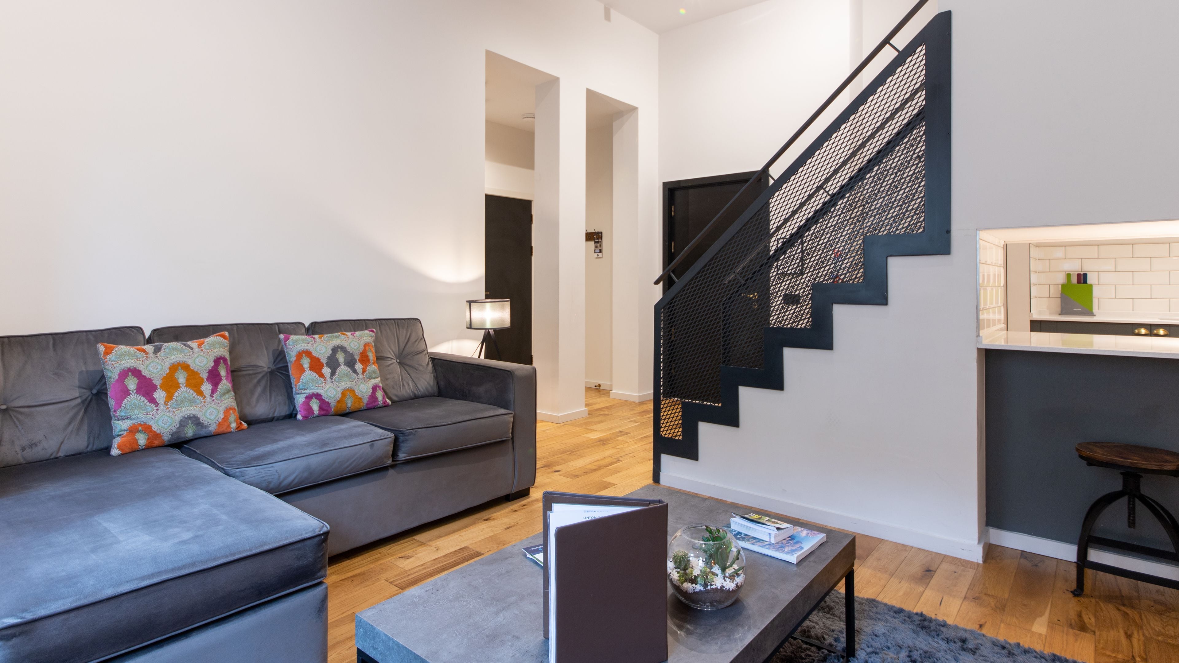 A modern living room with a grey sofa, colorful cushions, a coffee table, and a staircase with a black railing.