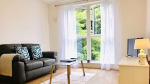 A living room with a sofa, a glass coffee table, a white side table with a lamp, and a large window with a view of greenery. - SFA New Central Woking (Woking)