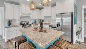 A kitchen with white cabinets, a granite island, stainless steel appliances, wicker stools, and pendant lighting.