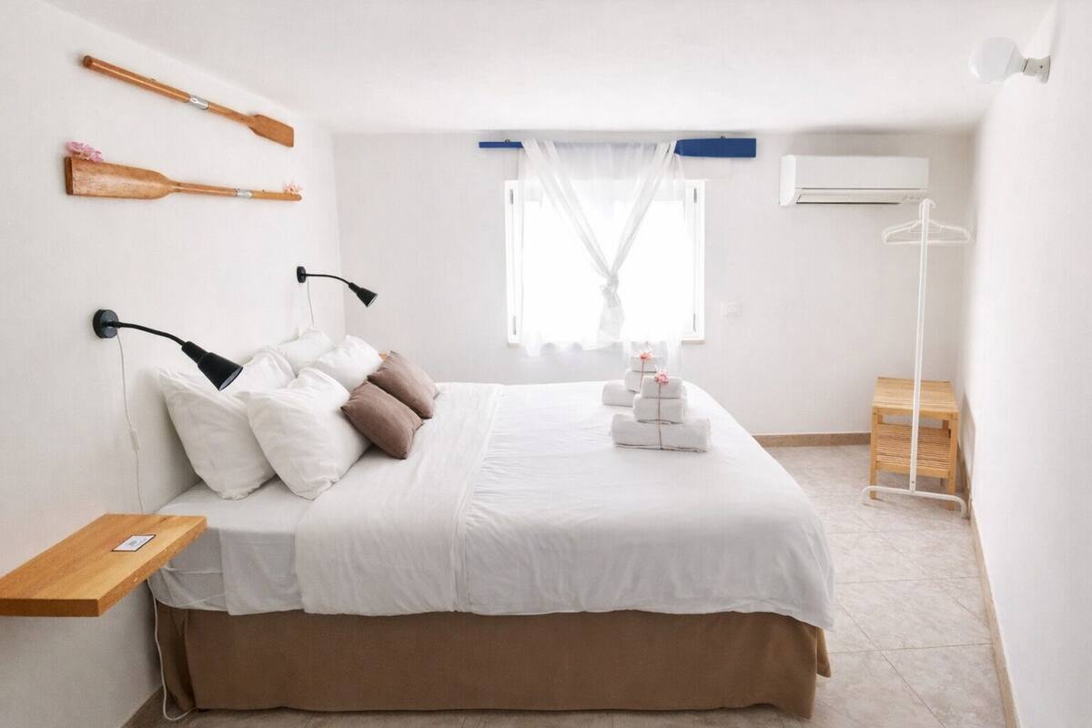 A bedroom with a bed, pillows, a bedside table, a hanging lamp, a window with sheer curtains, and a wooden floor.