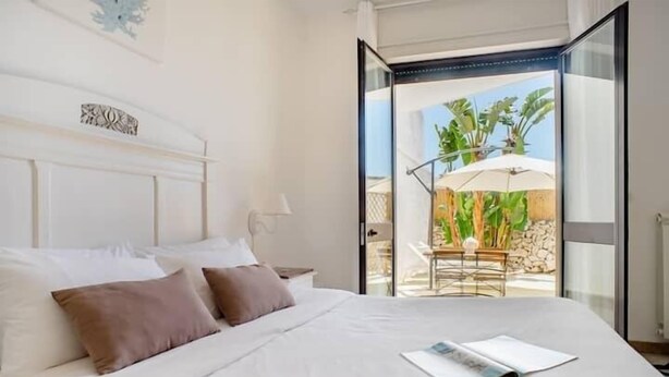 A bedroom with a large bed, a nightstand, and a window opening to an outdoor area with a table and umbrella. - Biancaleuca Rooms & Suite (Castrignano del Capo)