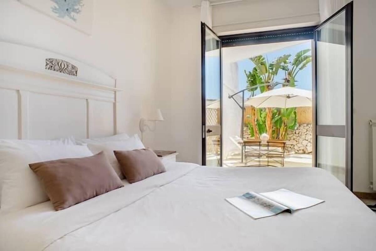 A bedroom with a large bed, a nightstand, and a window opening to an outdoor area with a table and umbrella.