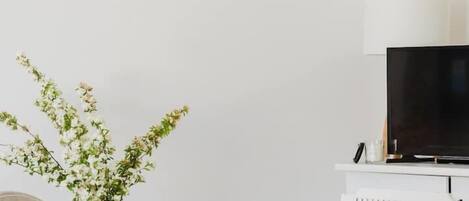 Un comedor con una mesa de madera, sillas blancas, un jarrón blanco con flores, una tetera con lunares, un televisor sobre un mueble blanco y un cuadro de una escena costera colgado en la pared.
