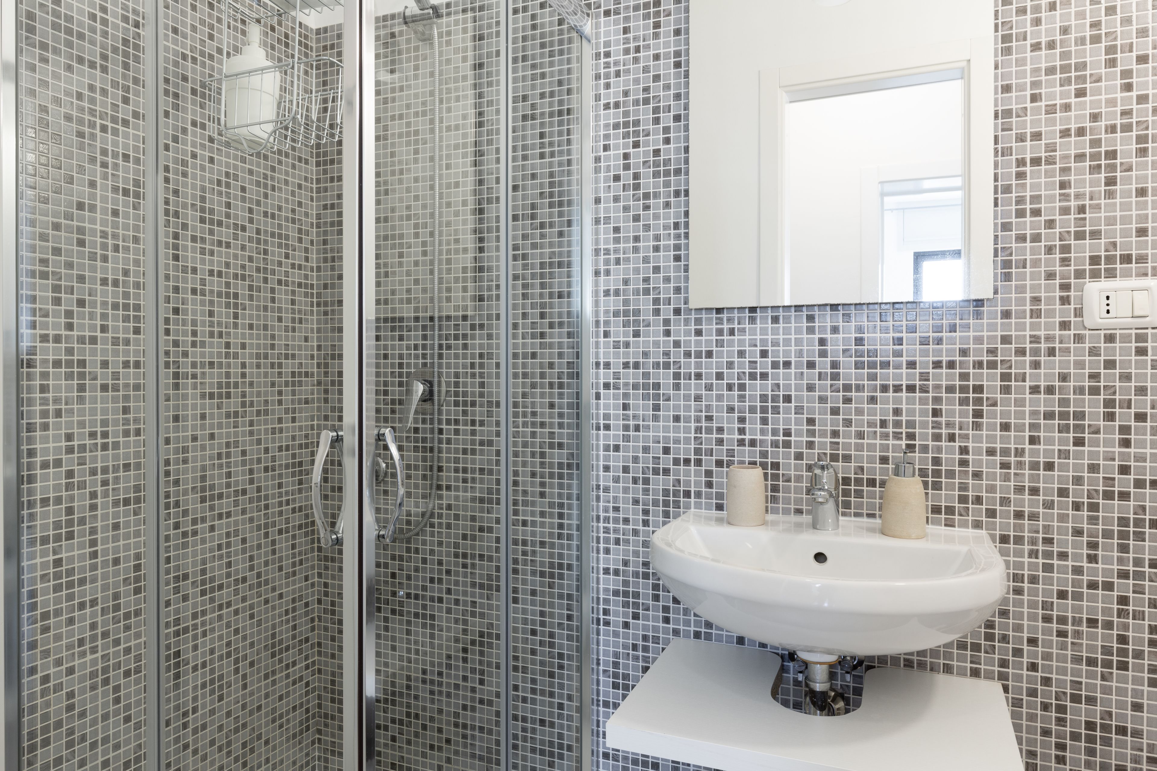 Une salle de bain avec une douche à parois de verre, un lavabo blanc avec un miroir au-dessus et un mur recouvert de carreaux.