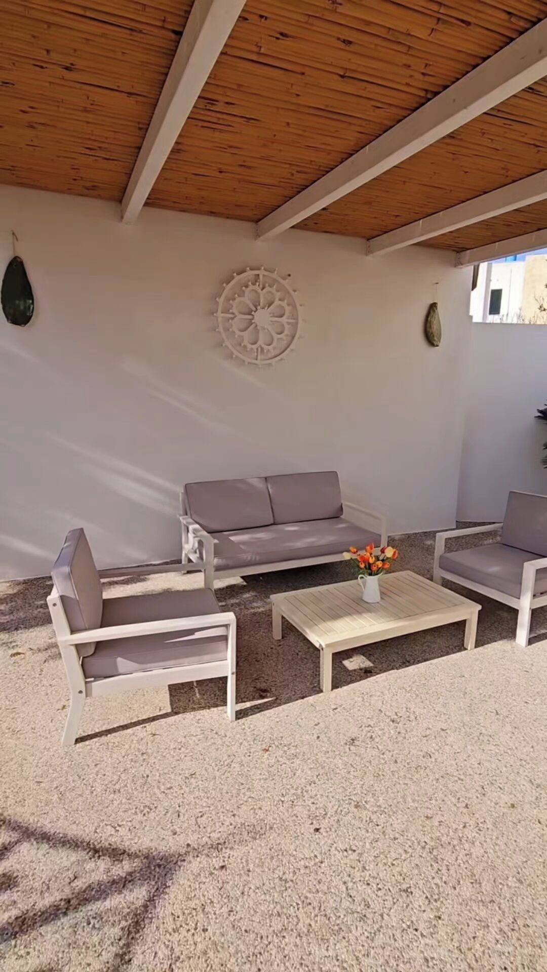 Un área de estar al aire libre con techo de madera, pared blanca con un adorno de rueda y sofá gris.