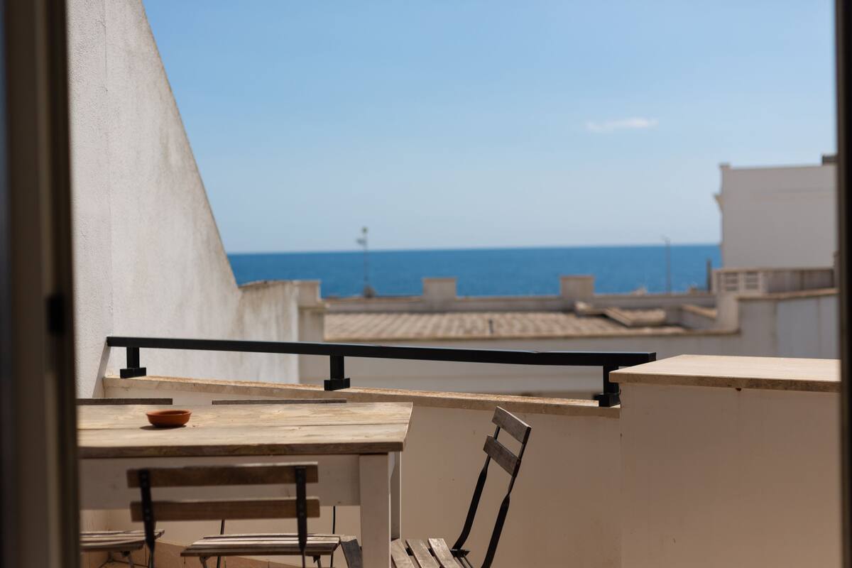 A balcony with a table and chairs, overlooking a coastal view.