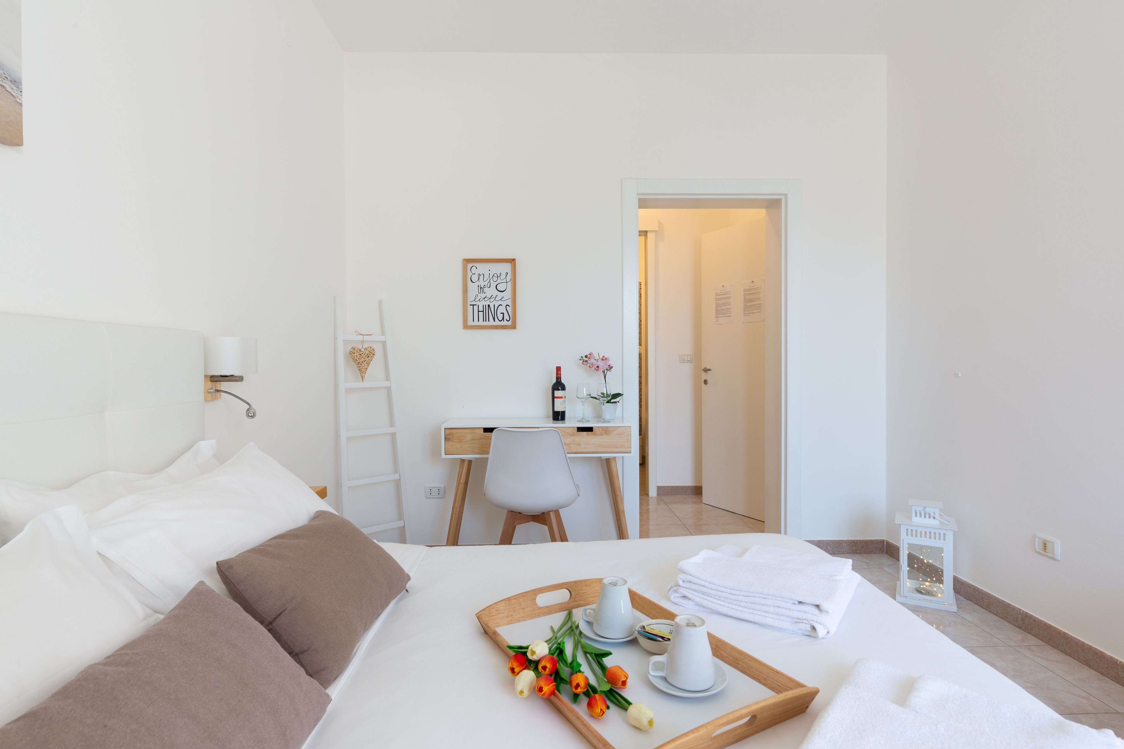 A hotel room with a bed, a desk, a chair, a tray with flowers, and a small table with a cup and a bowl.