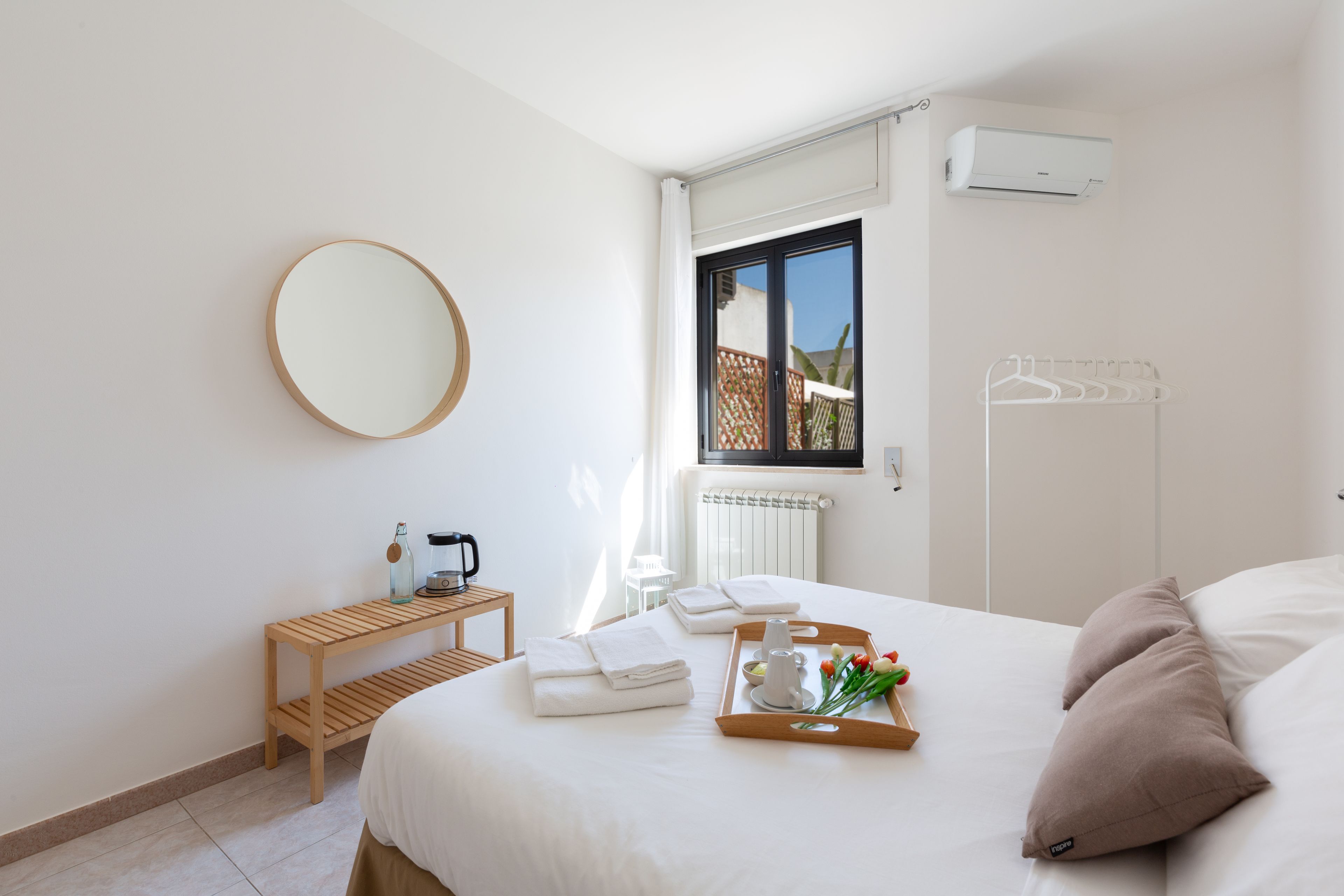 A hotel room with a bed, a wooden side table with a kettle, a tray with towels and flowers, a round mirror, and a window with a view of a fence and trees.