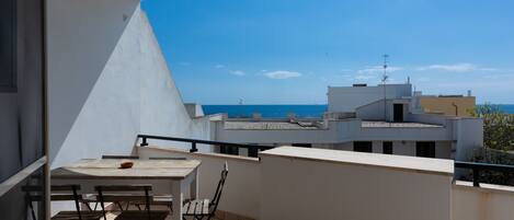 A balcony with a table and chairs, overlooking a coastal area with buildings and the sea.
