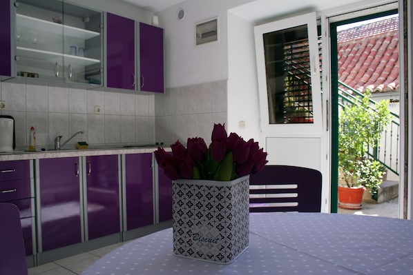Eine Küche mit violetten Schränken, einer gefliesten Spritzschutzwand und einem Fenster mit Blick auf ein rotes Ziegeldach und Grünflächen.