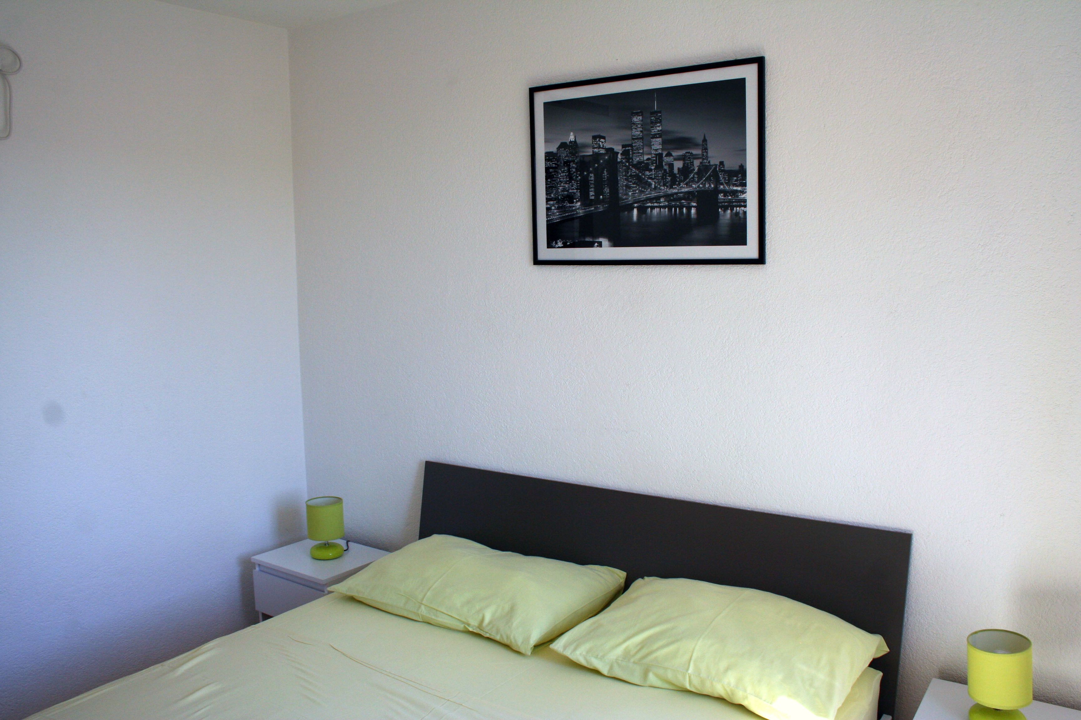 A bedroom with a bed, two pillows, a nightstand, a lamp, and a framed picture on the wall.