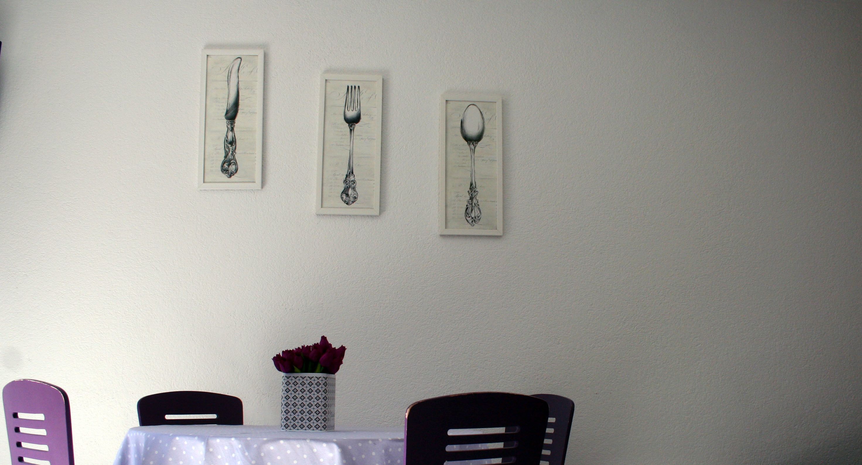 Un comedor con mesa y sillas, un jarrón con flores y tres utensilios enmarcados en la pared.