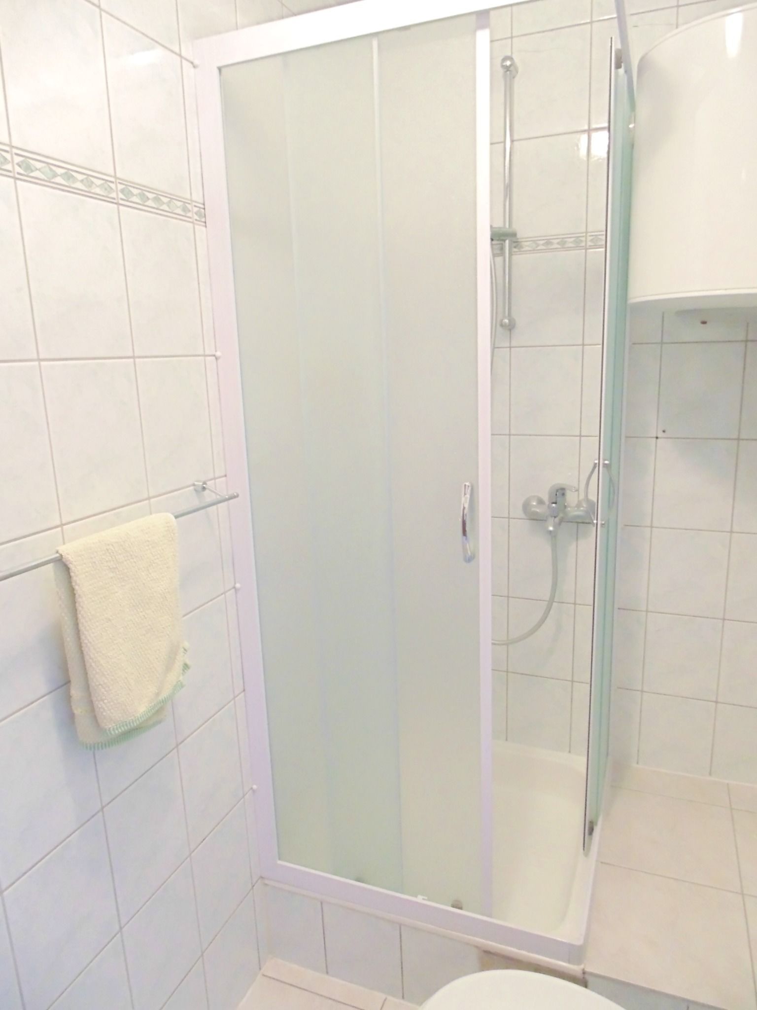 A glass-enclosed shower stall with a white tiled wall and a towel hanging on a rack.