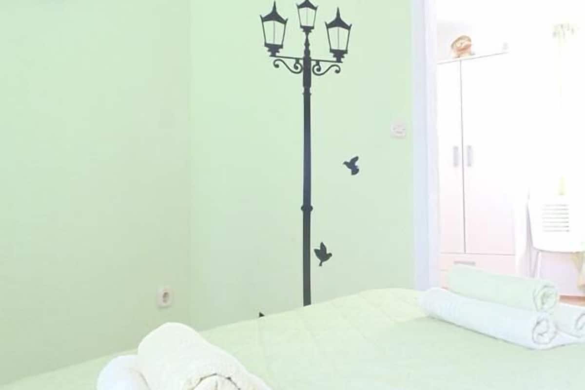 A hotel room with a bed, a wall-mounted air conditioner, a wall-mounted lamp, and a view of the outside.