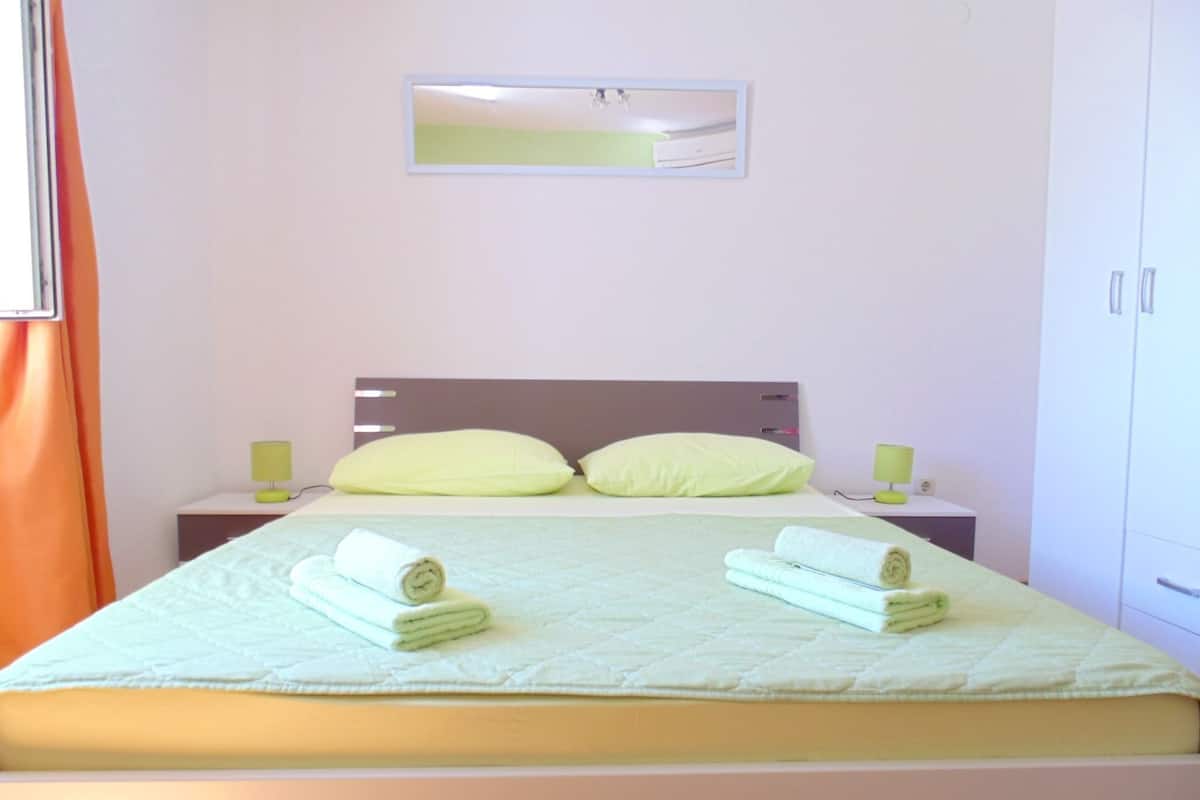 A neatly made bed with two pillows and two rolled towels, a bedside table with a lamp, and a mirror on the wall.
