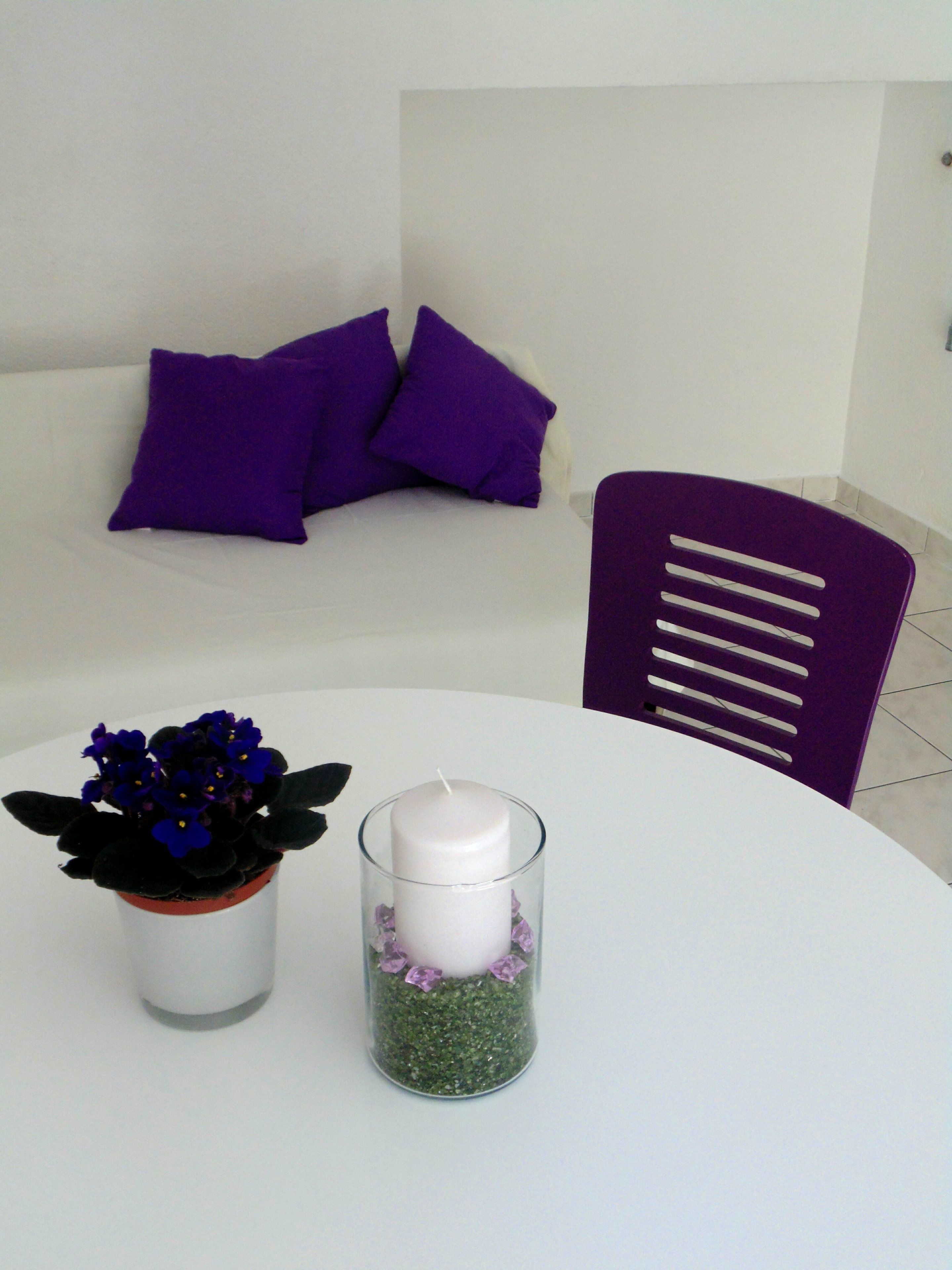 Una habitación con un sofá blanco, almohadas moradas, un sillón morado, una mesa blanca con un candelabro de cristal y una planta en maceta.