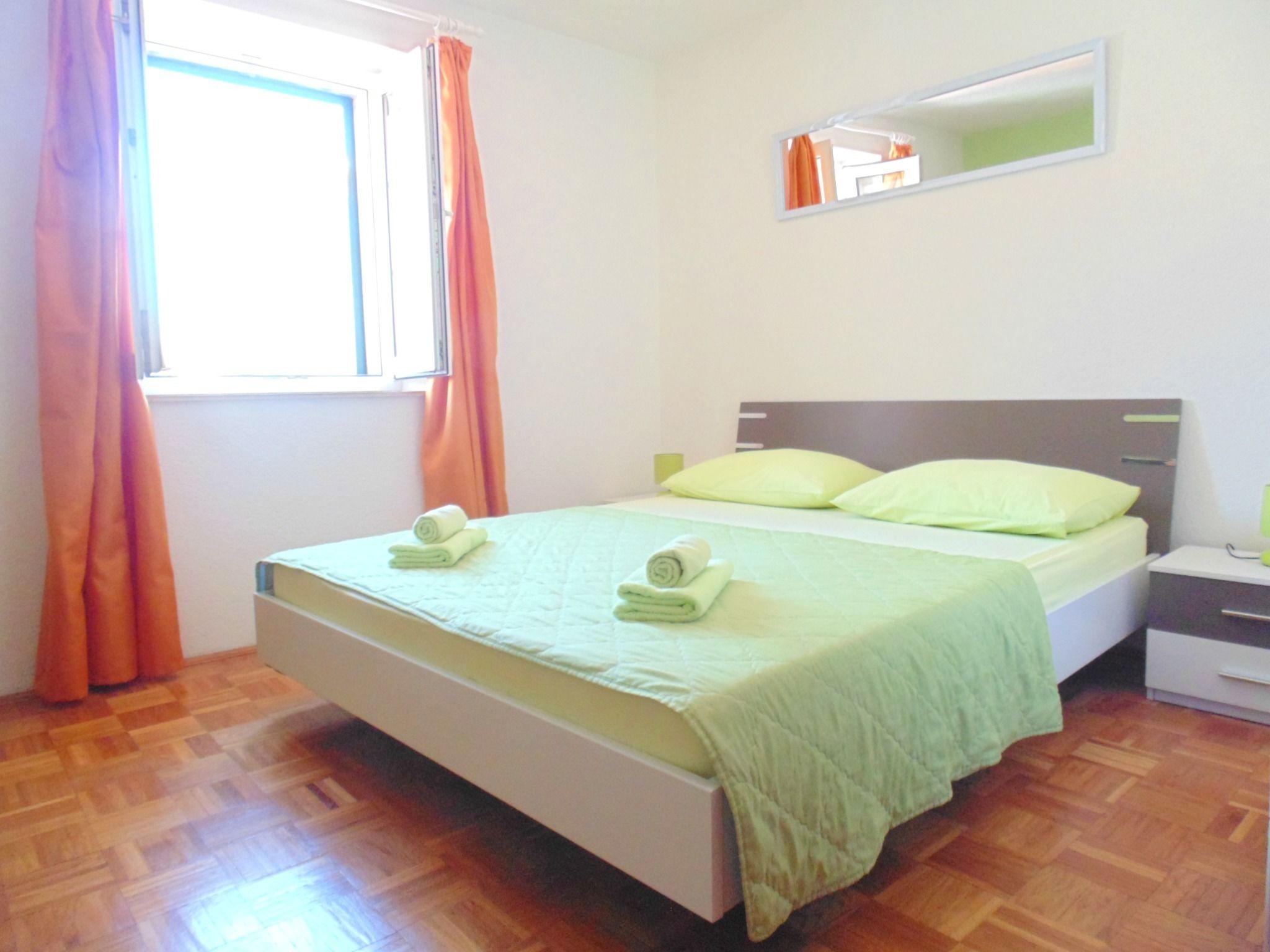 Un letto ben rifatto con copriletto e cuscini verdi, pavimento in legno, un comodino con una lampada e una finestra con tende arancioni.