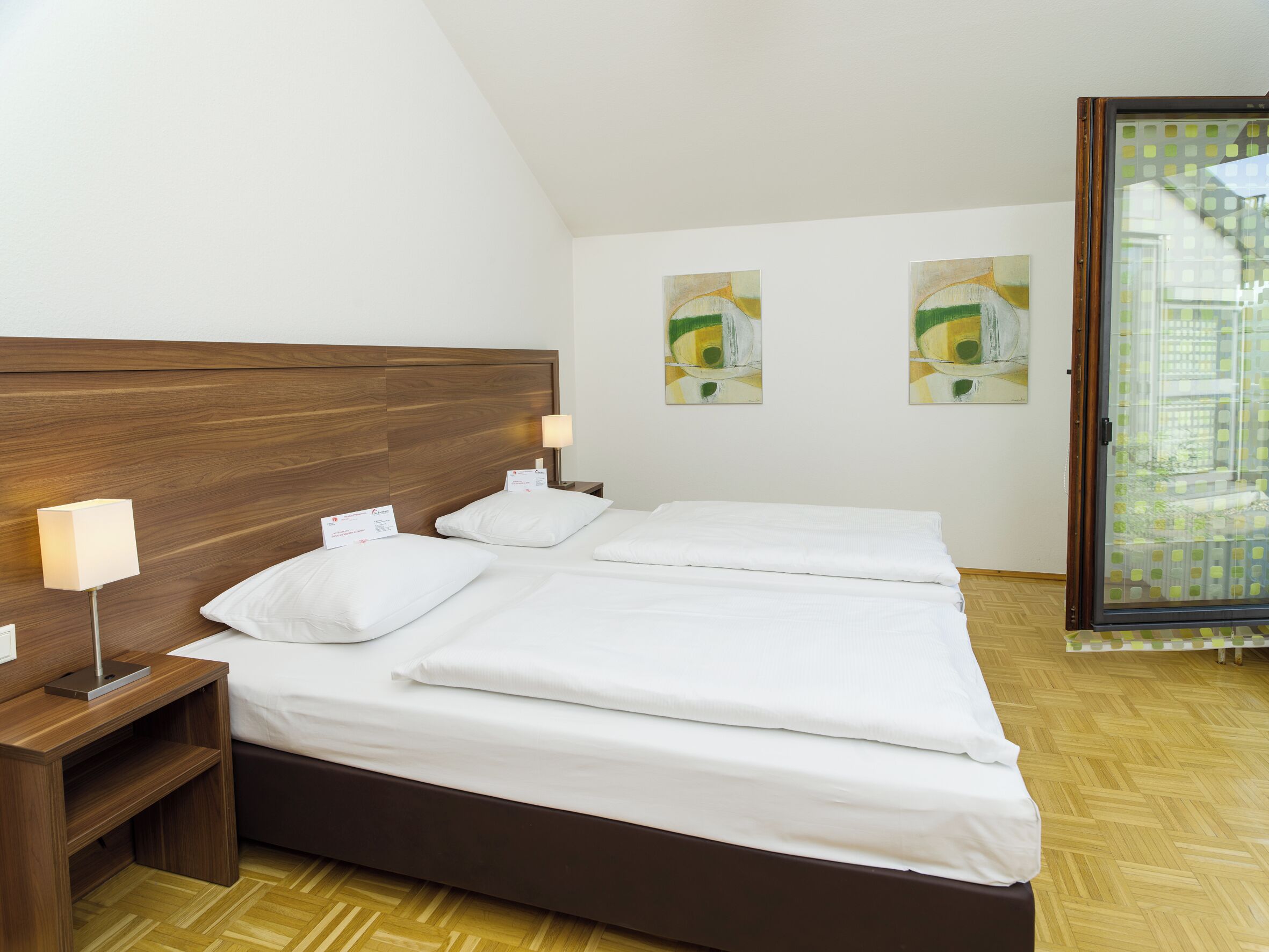 Ein Hotelzimmer mit einem Holzkopfteil, einem Bett mit weißen Leinentüchern, einem Nachttisch mit einer Lampe und zwei abstrakten Bildern an der Wand.