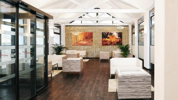Un interior moderno con paredes de vidrio, piso de madera y una zona de estar central con sofás blancos y un tapete estampado.