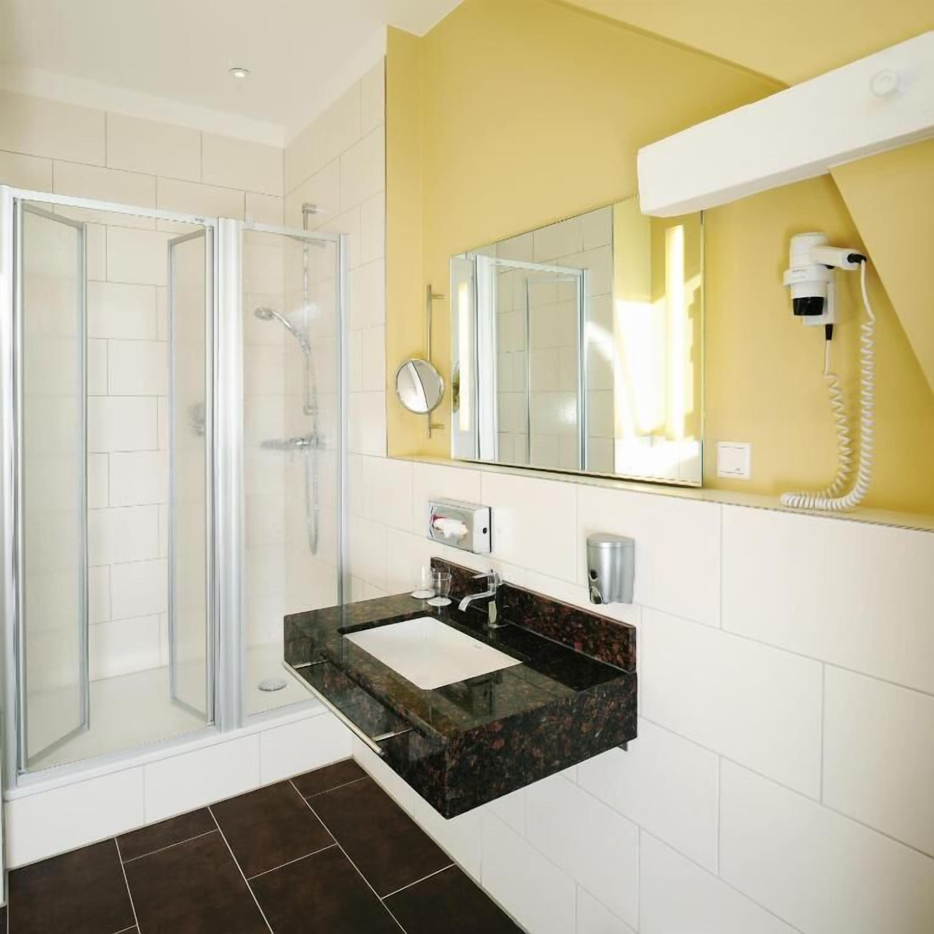 A bathroom with a glass shower enclosure, a sink with a marble countertop, a mirror, and a hairdryer mounted on the wall.
