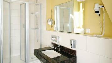 A bathroom with a glass shower enclosure, a sink with a marble countertop, a mirror, and a hairdryer mounted on the wall.
