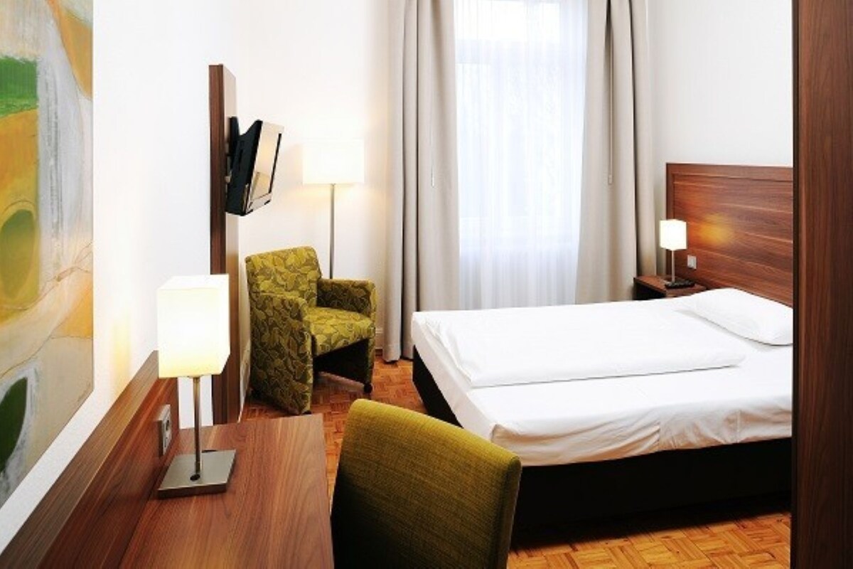 Habitación de hotel con cama, una silla, escritorio, lámpara y un cuadro en la pared.