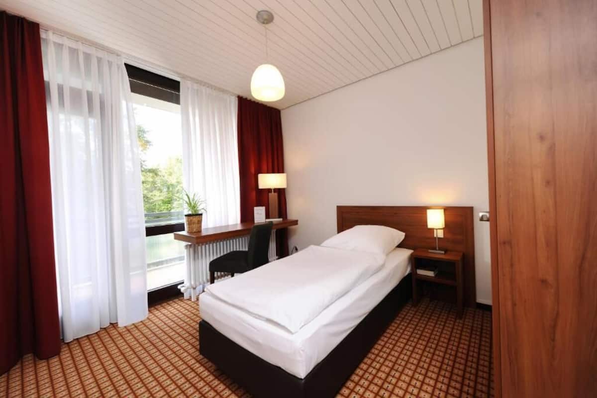 Habitación de hotel con cama, mesitas de noche, escritorio, silla y ventana con cortinas.