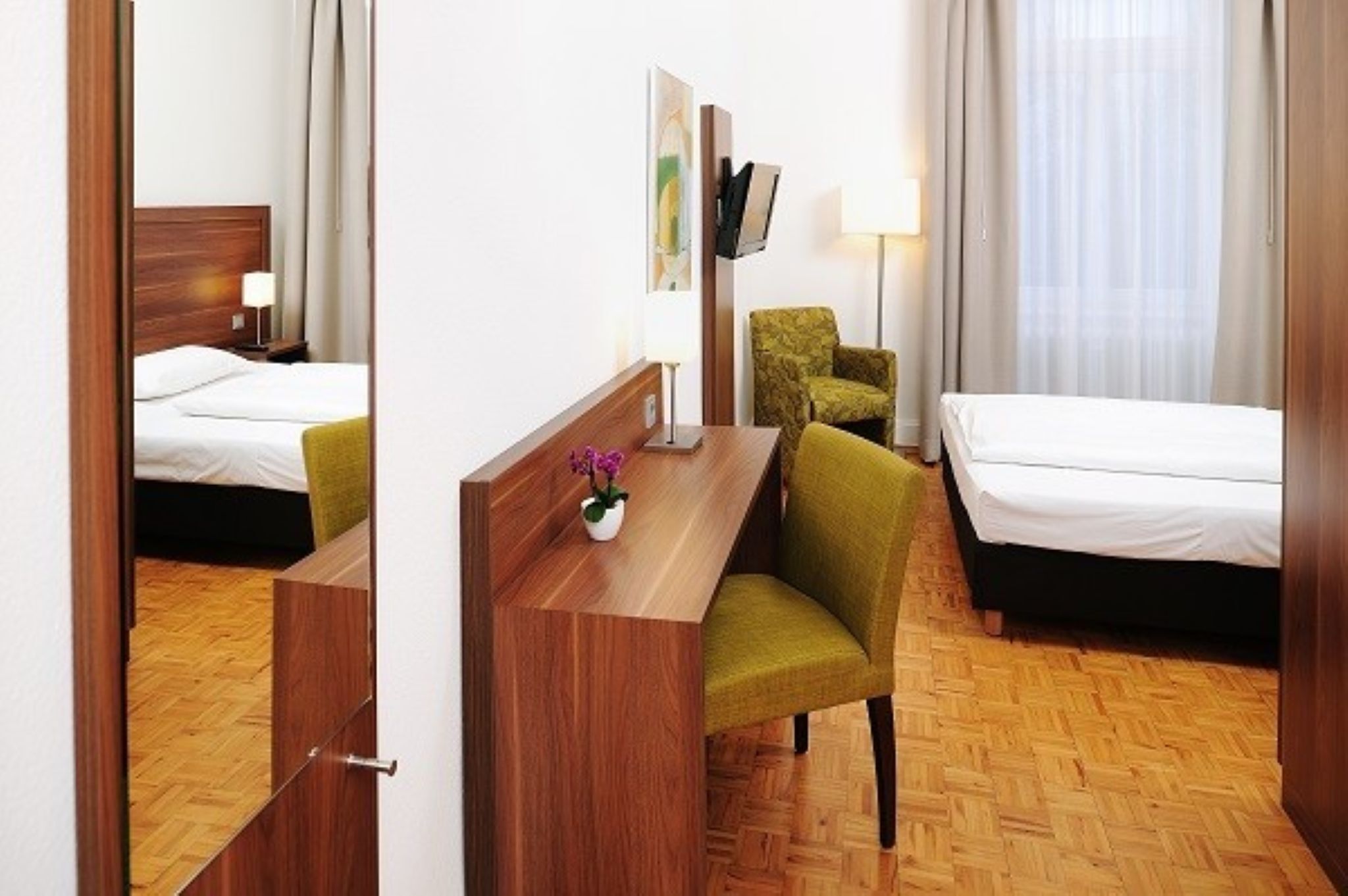 Habitación de hotel con un escritorio de madera, dos camas, una silla verde, un televisor y una lámpara.
