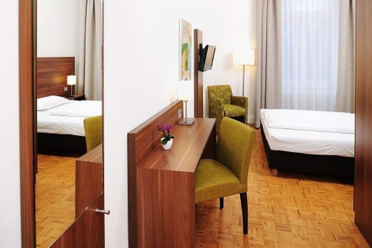Habitación de hotel con un escritorio de madera, dos camas, una silla verde, un televisor y una lámpara.