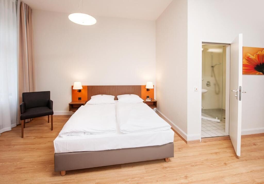 A hotel room with a double bed, two bedside tables with lamps, a chair, and a door leading to a bathroom.