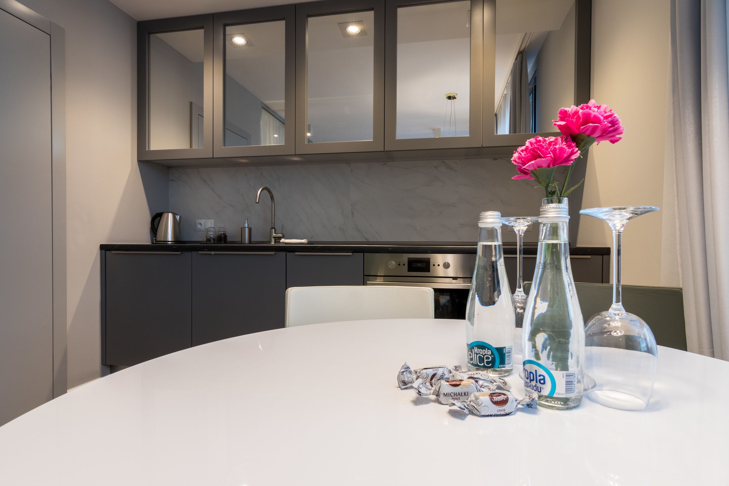 Una cocina moderna con armarios oscuros, una encimera blanca y una mesa de comedor blanca con un jarrón de flores y botellas.